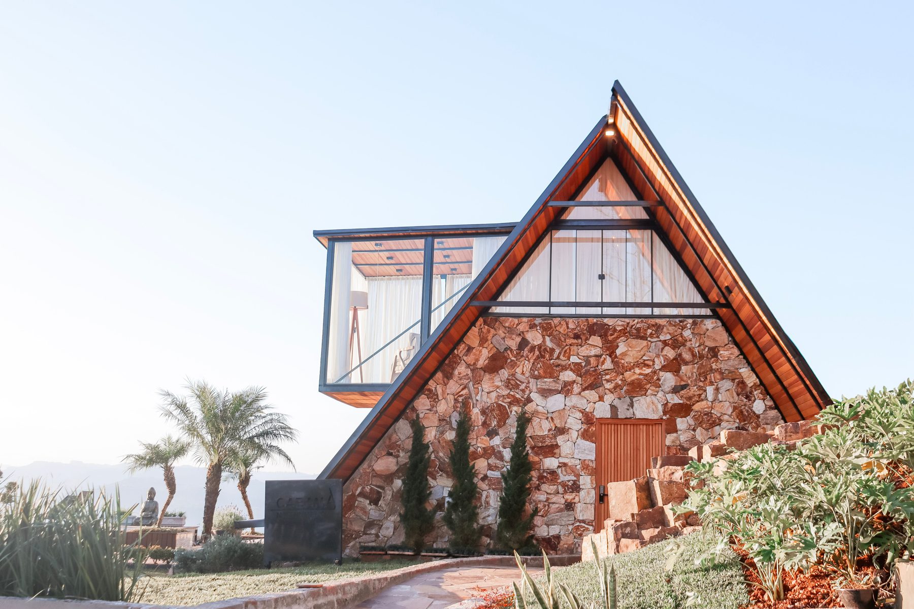 Fachada triangular da cabana com pedra, vidro e jardim